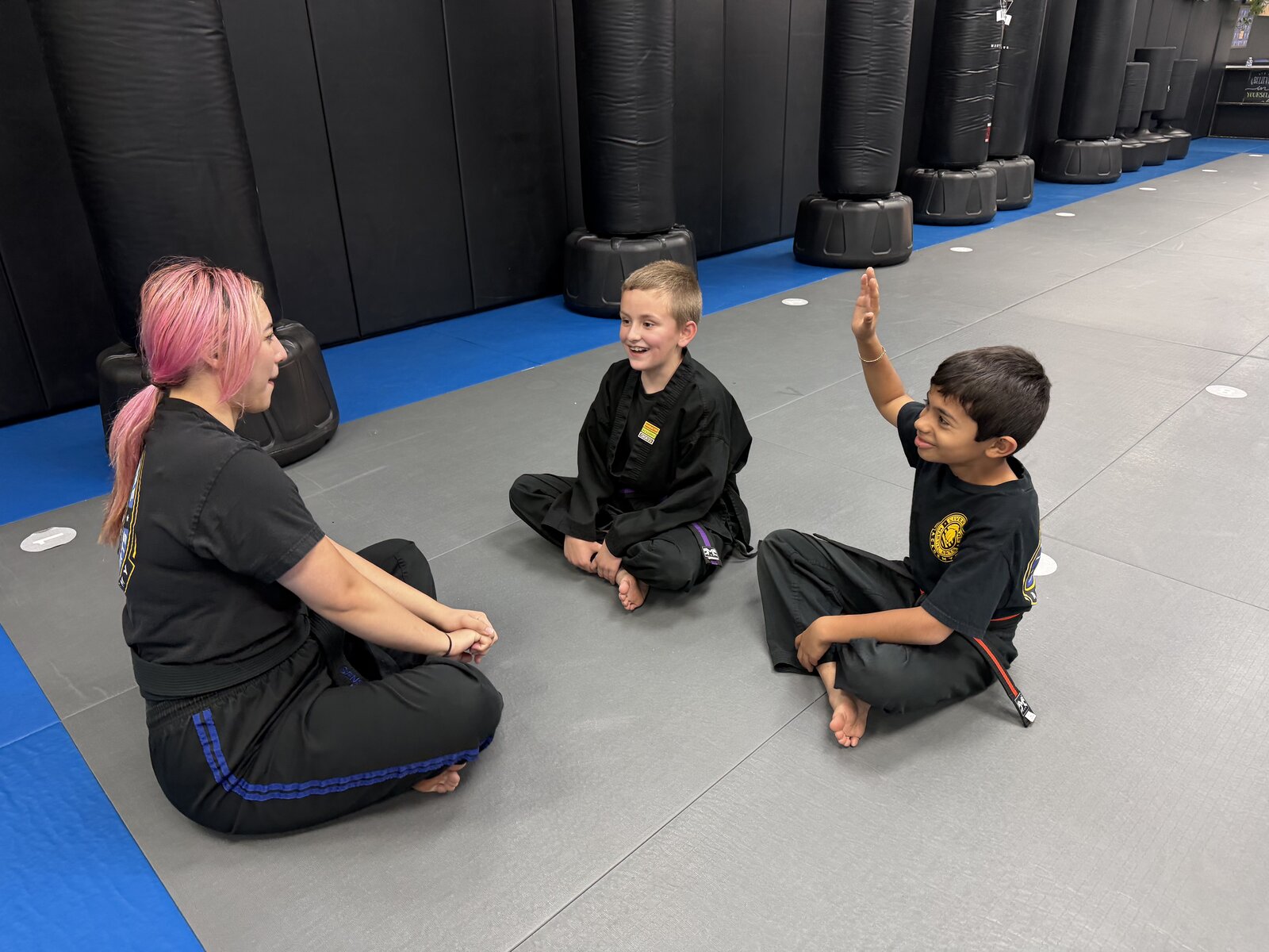 An instructor sitting on the mats with two young students, one raising his hand, at Livermore Martial Arts Academy